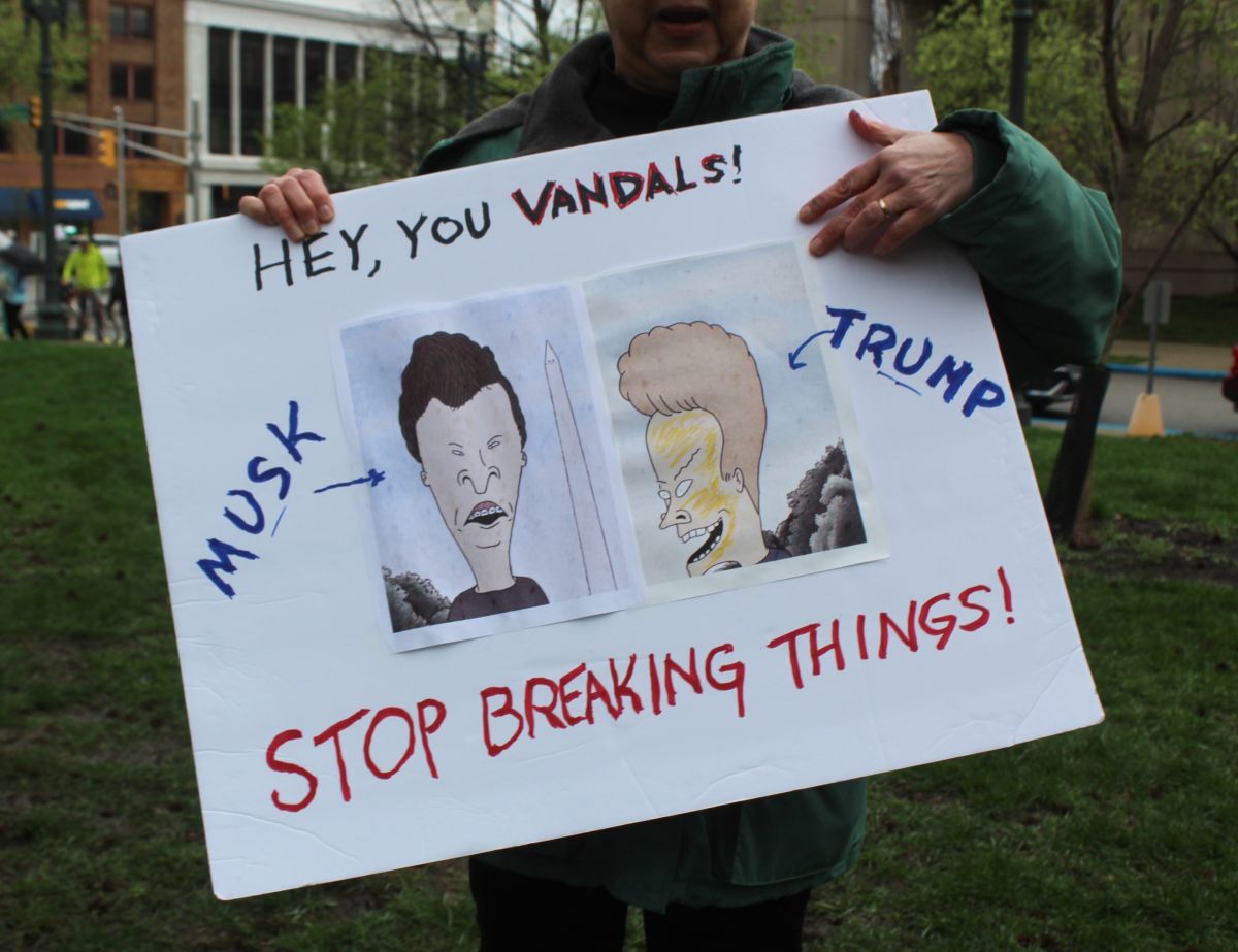 Julie Stahlhut, a Beech Grove resident and retired science researcher, holds a handmade sign with comical depictions of Elon Musk and Donald Trump. “I wanted to have some humor in it, because this is a serious subject,” she said. Stahlhut and her husband, both retired, say they’ve watched their savings dwindle as Trump’s tariffs have shaken the stock market.