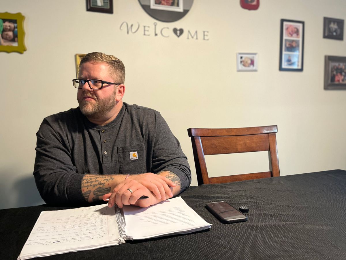 A man sitting at his dining room table in front of a wall with family photos and the word welcome.