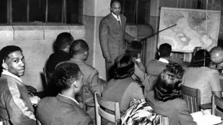 Students sit at their desks at Crispus Attucks High School in 1948 and face the front of the classroom where the teacher is pointing at a map. - Indianapolis Recorder Collection, Indiana Historical Society