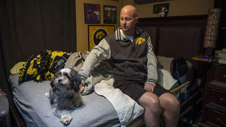 Jon Miller sits in his bedroom with his dog, Carlos, who he received as a present for successfully completing cancer treatment a decade ago. Miller sustained severe brain damage from advanced testicular cancer and its treatment. He requires the help of home health aides to continue living in his West Des Moines home. - Natalie Krebs / Side Effects Public Media