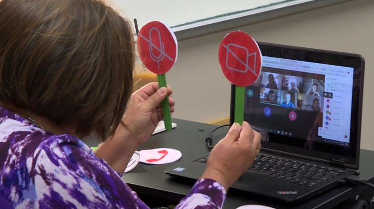 Tami McMahan sits in front of a computer on the first day of school, holding up icons representing the microphone and webcam buttons to help her virtual third grade class see whether they should turn theirs on or off.  - Jeanie Lindsay/IPB News