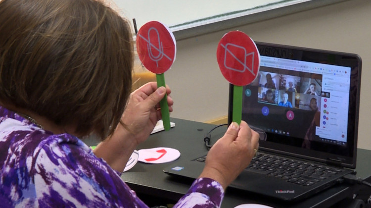 Tami McMahan sits in front of a computer on the first day of school, holding up icons representing the microphone and webcam buttons to help her virtual third grade class see whether they should turn theirs on or off. - Jeanie Lindsay/IPB News
